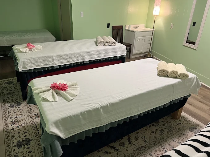 Spacious couples massage room at Golden Hands Massage featuring clean therapy side-by-side beds with fresh white linens, decorative towel art with pink flowers, soft lighting, and calming pale green walls.